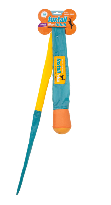 The Foxtail Fetch Dog Toy features a bright orange and yellow ball with a long blue and yellow fabric tail, designed for slobber-free play and shown in its packaging.