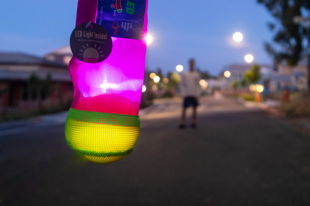 The Foxtail LED glows pink and yellow in the foreground, resembling a light-up night toy, while a person stands blurred in the background on an empty street at dusk.